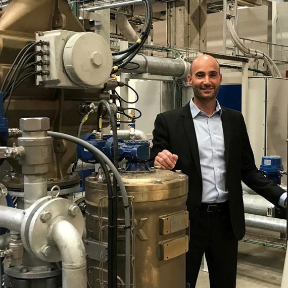 Man in industrial setting with large machinery and pipes in the background.