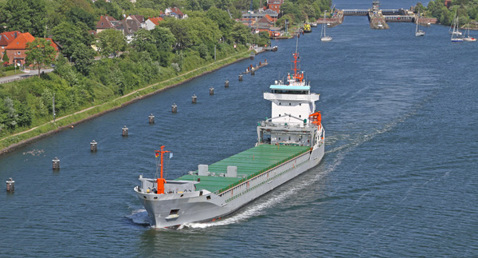 Cargo ship navigating a canal near a town with lush greenery.