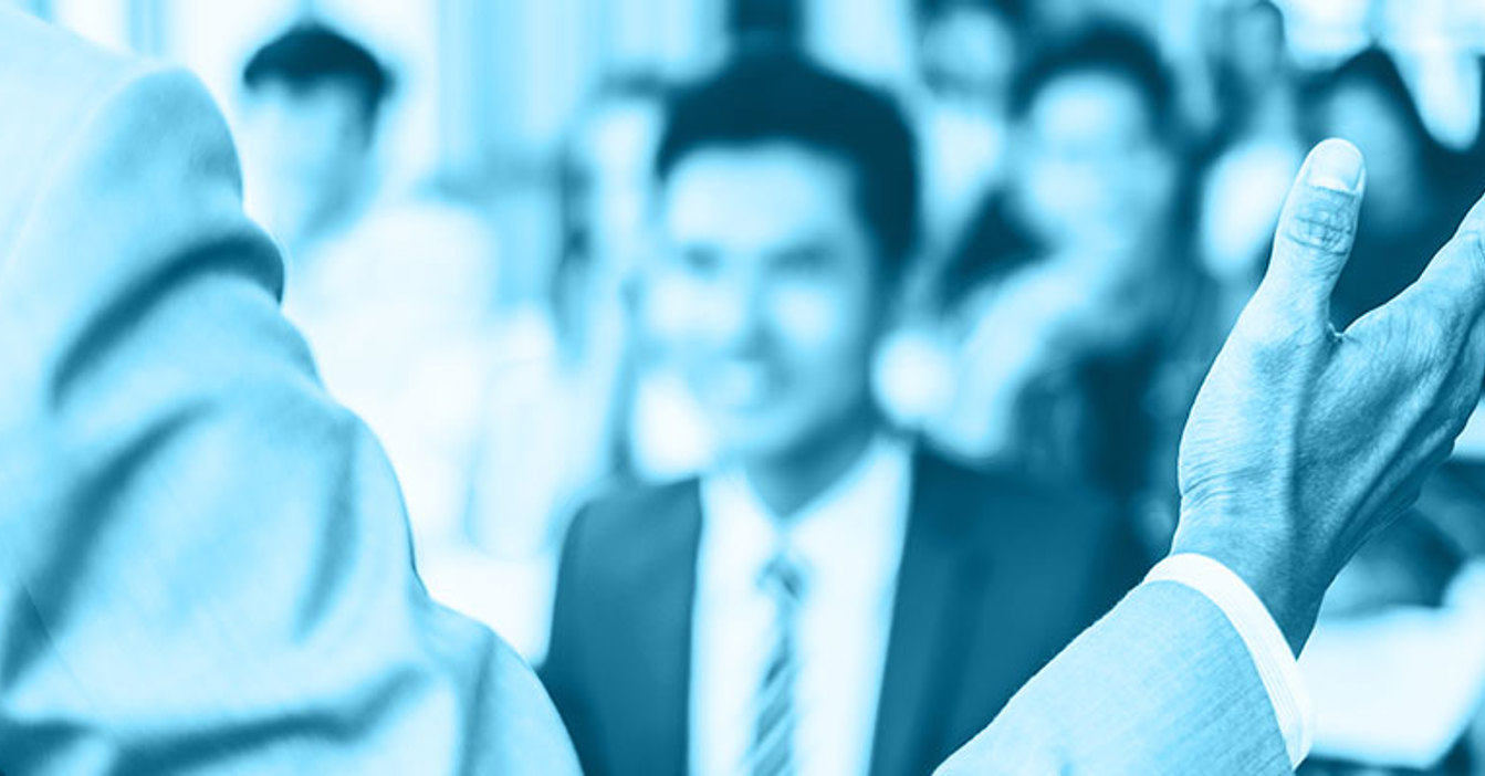 Person in a suit gesturing during a presentation to a blurred audience.