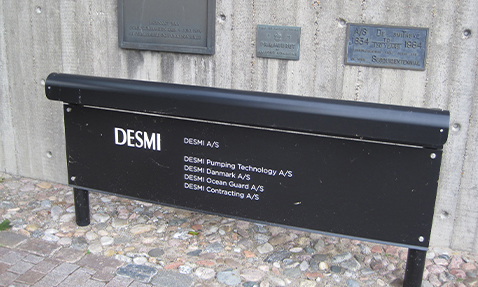 Black sign displaying various DESMI company names on a stone wall background.