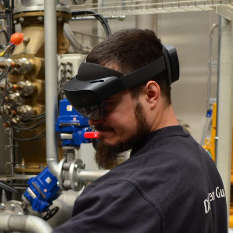 Technician wearing augmented reality headset in industrial facility with pipes and machinery.
