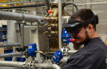 Technician wearing augmented reality headset in industrial facility with pipes and machinery.