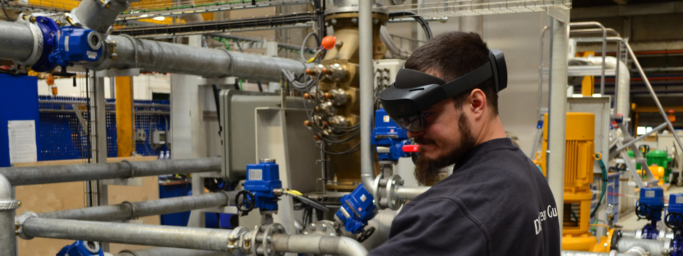 Technician wearing augmented reality headset in industrial facility with pipes and machinery.