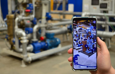 Hand holding smartphone with augmented reality view of industrial pipes and machinery.