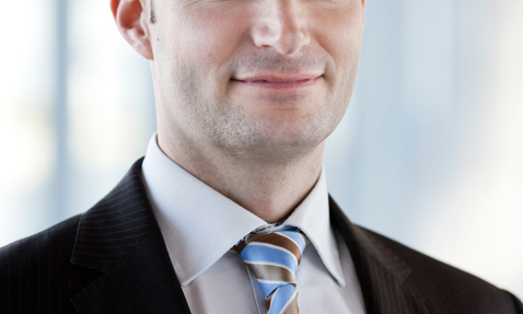Smiling man in suit and striped tie, standing indoors with blurred background.
