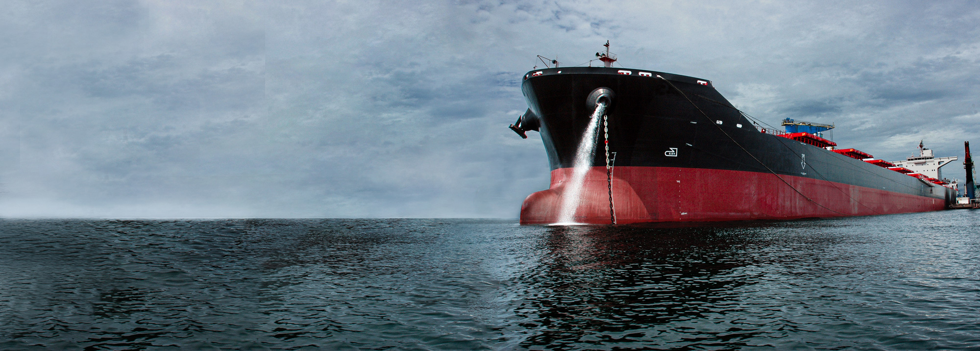 Large cargo ship docked, releasing water from the bow, under cloudy skies.