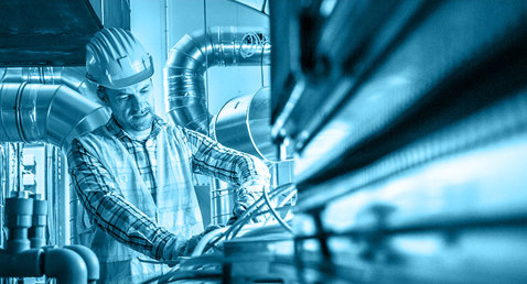 Worker in hard hat inspecting industrial pipes and machinery in a facility.