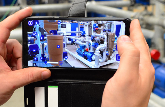 Hands holding a smartphone displaying augmented reality of industrial pipes and machinery.