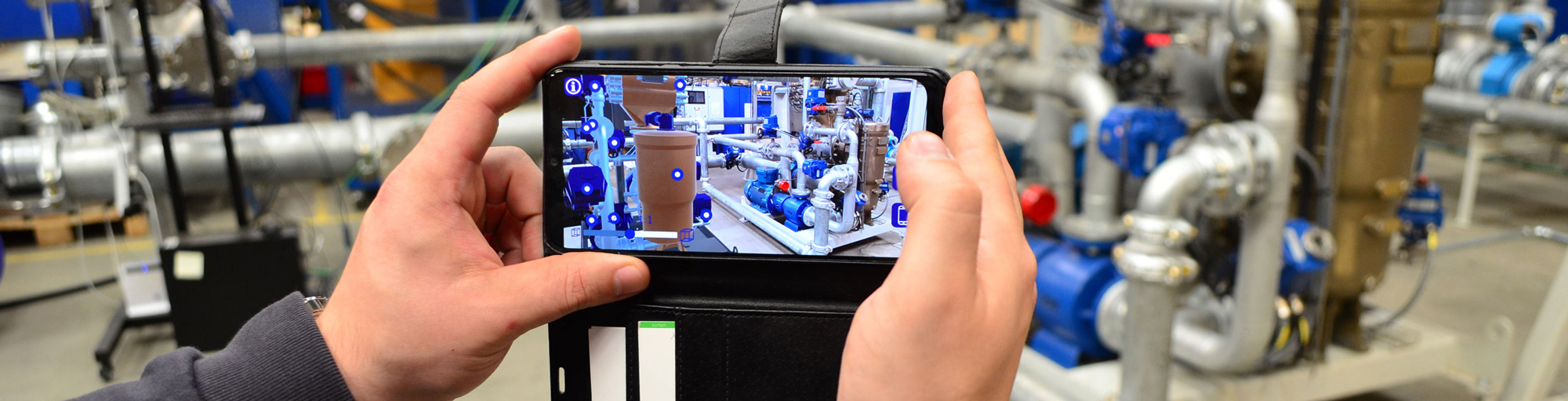 Hands holding a smartphone displaying augmented reality of industrial pipes and machinery.